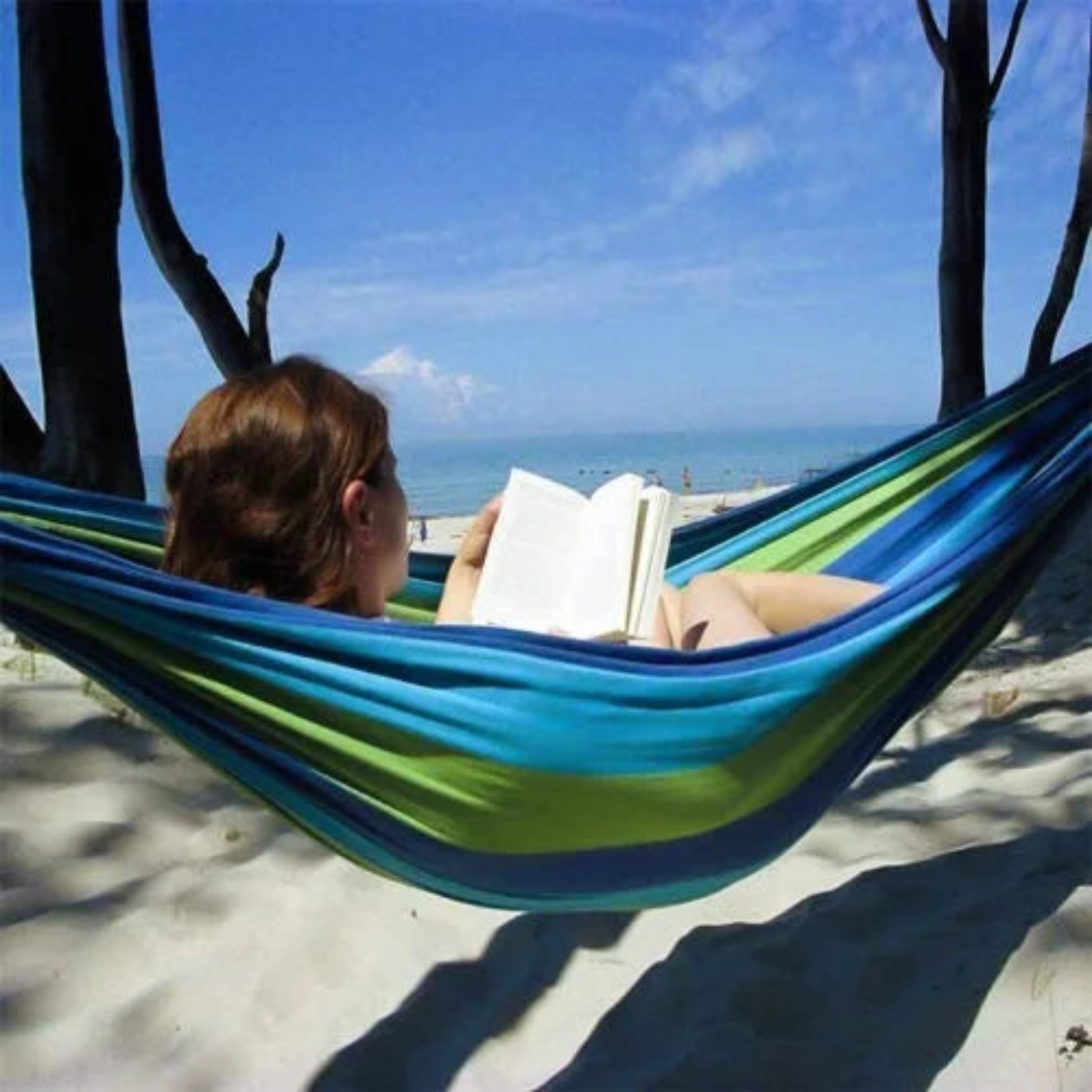 Person reading a book in a blue and green hammock on a beach.